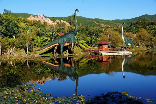 Life-size sauropod sculptures reflected in a calm pond at Lufeng World Dinosaur Valley, Yunnan, China.