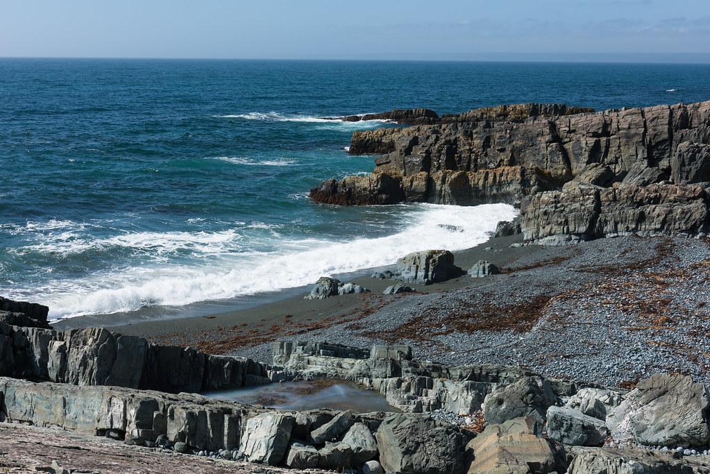 Grey sea cliffs and layered rocky tidal platforms with surf rolling onshore at Mistaken Point, Newfoundland.