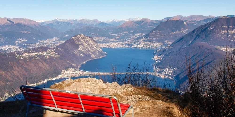 Red bench on a mountain ridge overlooking Lake Lugano with distant snow-capped peaks above Monte San Giorgio.