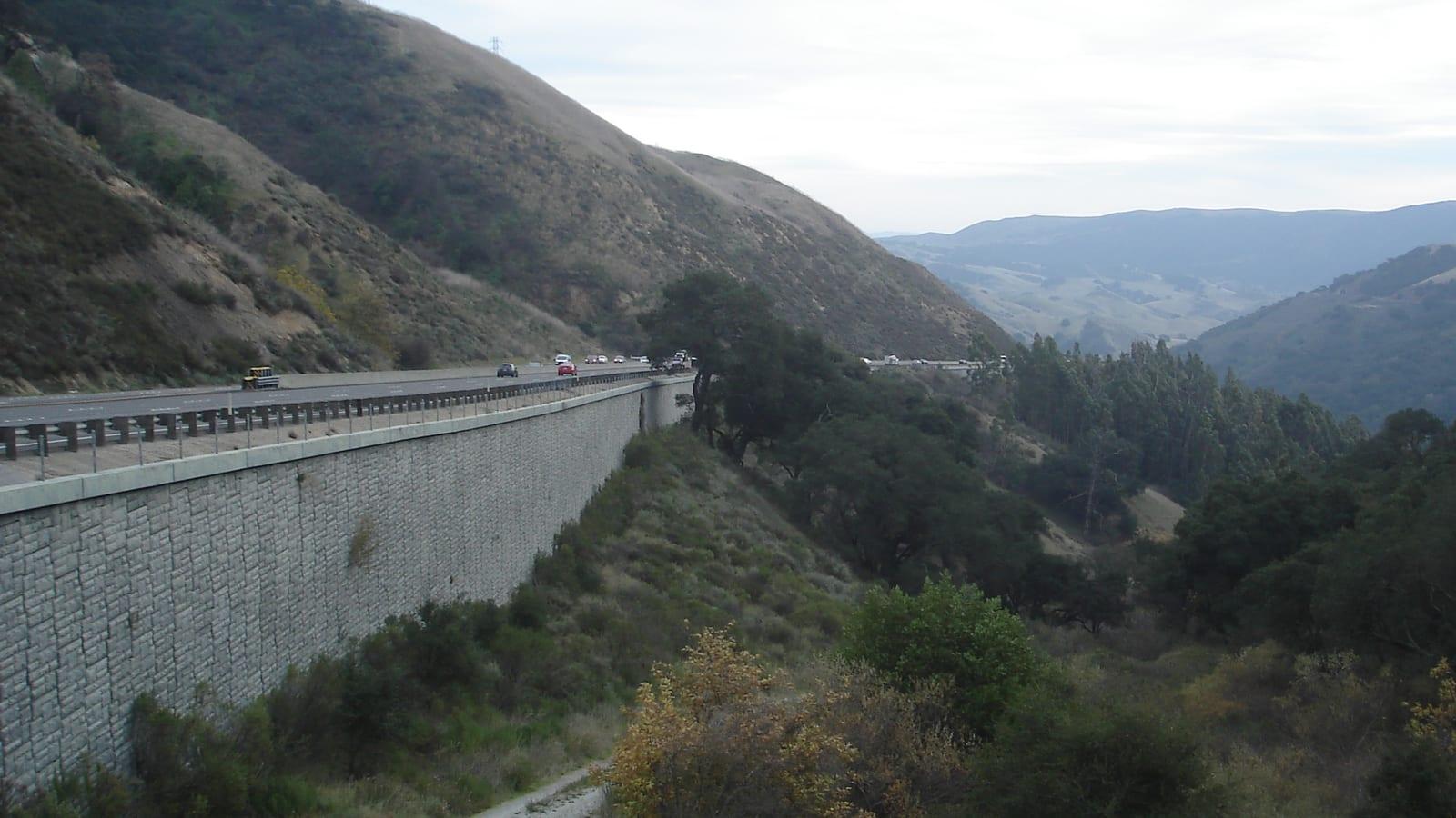 Highway with stone retaining wall climbing between grassy hills and oaks near Santa Margarita Ranch, California.
