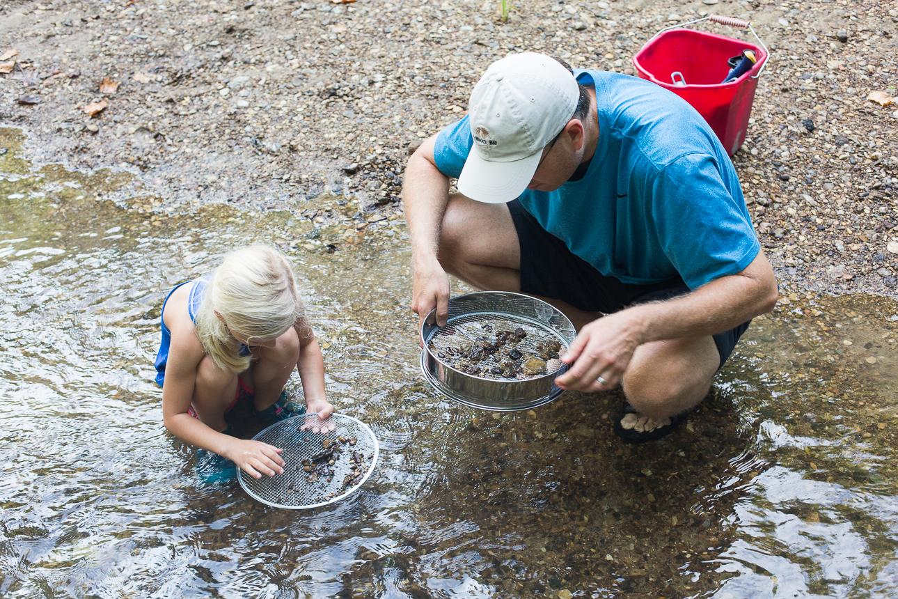 Adult and child sieving gravel in the shallows of a creek with a red bucket on the bank at Post Oak Creek, Texas.